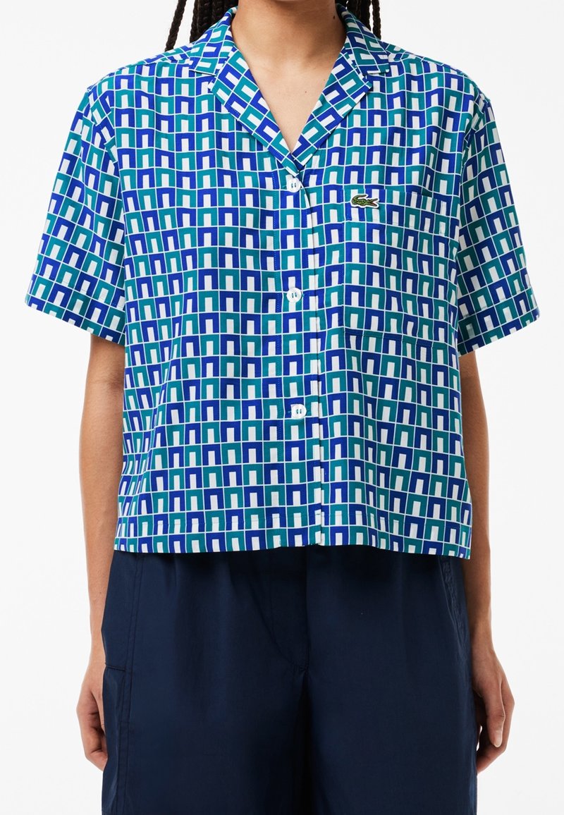 Chemise à manches courtes boutonnée avec un motif géométrique bleu, vert et blanc ; elle possède un col et un petit logo sur la poitrine.