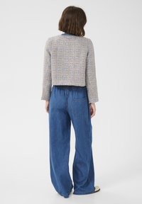Cropped, textured, beige jacket with light blue accents over loose, blue denim trousers featuring a back pocket and elastic waistband.