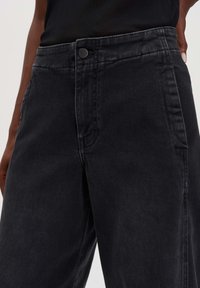 Schwarze Jeans mit hoher Taille, die über einen vorderen Knopfverschluss und seitliche Taschen verfügt. Der Stoff hat eine glatte Textur und eine leichte Ausbleichung.