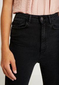 Mustat farkut, joissa on korkea vyötärö, viisi taskua, metallinen nappikiinnitys ja hienovarainen rakenne. Kädessä näytetään kokovertailuksi.