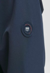 Navyblauer Stoff mit einem kreisförmigen schwarzen Aufnäher, auf dem in rotem und weißem Text "JACK & JONES" und "JDJ DENIM SOCIETY" steht. Glatte Textur.