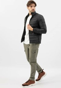 Giacca nera imbottita con colletto alto, t-shirt bianca, pantaloni cargo olive con tasche e scarpe alte marroni con suole bianche.