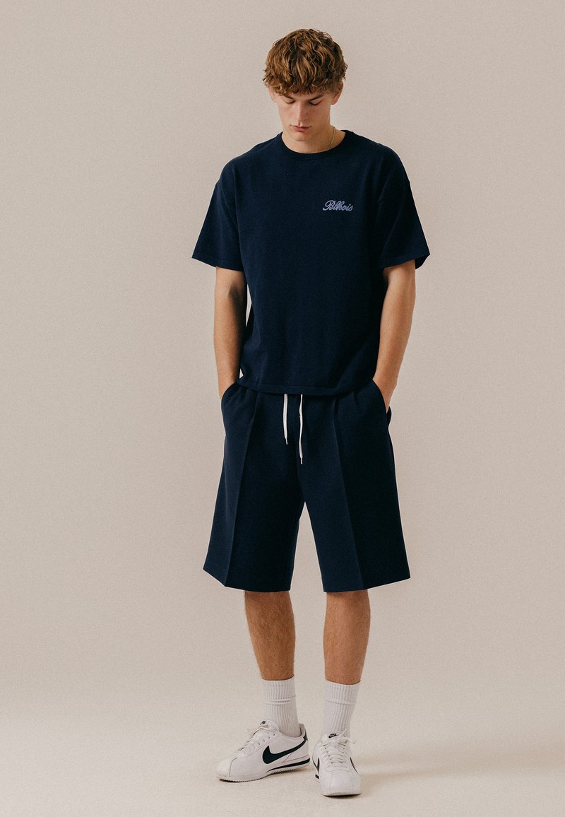Jeune homme portant un t-shirt bleu marine et un short, des chaussettes blanches et des baskets blanches Nike, debout les mains dans les poches et regardant vers le bas.