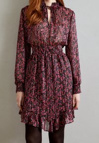 Abito floreale nero con maniche lunghe, fascia arricciata in vita, orlo con volant e scollatura con fiocco. Presenta motivi di fiori rosa e viola su tessuto trasparente.