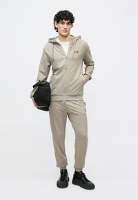 Beige hettejakke med glidelås og matchende bukser i mykt stoff, med en logo på brystet, kombinert med svarte joggesko og en svart veske.