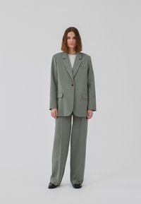 Lys grønn skreddersydd dress med en knapp jakke, vide bukser og en subtil lomme detalj. Brukt over en lys grå topp.