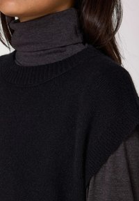 Maglione a collo alto in tessuto di maglia nero con motivo testurizzato, ampio colletto a coste e indumento grigio stratificato sotto.