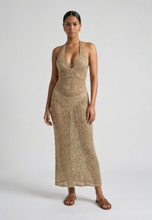 Donna che indossa un vestito beige all'uncinetto senza maniche con una scollatura profonda e sandali marroni bassi, in piedi su uno sfondo semplice.