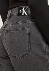 Tmavě šedé džíny vyrobené z výrazné texturované látky, s výrazným černým koženkovým štítkem na pase s logem značky.