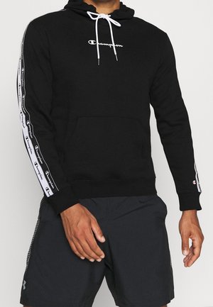 Mann trägt einen schwarzen Champion-Hoodie mit weißem Logo und Streifen auf den Ärmeln, kombiniert mit schwarzen Under Armour-Shorts.