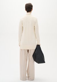 Beige lång cardigan med ribbat nederkant och manschetter, i kombination med ljusbeige vida byxor. Svart texturerad tote-väska som hålls i handen.