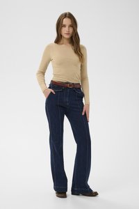 Bézs hosszú ujjú felső sötétkék, magasderekú bővülő farmerrel, amely kontrasztos fehér varrással és barna bőr övvel van ellátva.