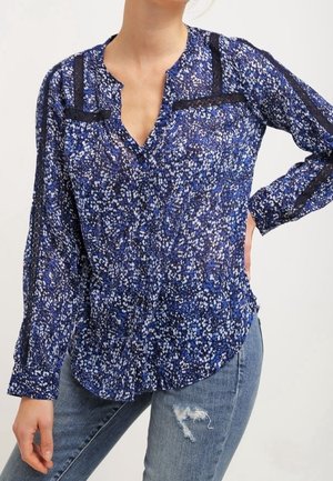 Kvinde iført en blå bluse med blomsterprint og sort detalje, kombineret med lyseblå revnede jeans, poserer med den ene hånd på hoften.