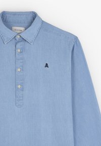 Camisa de botón azul claro hecha de algodón, con cuello, mangas largas y un pequeño logo de calavera negra en el lado izquierdo del pecho.
