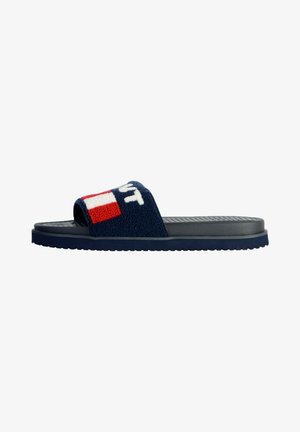 Navy slide sandal featuring a textured, fuzzy strap with a logo and red accents, and a flat rubber sole with a ridged pattern.