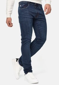 Mørkeblå denimjeans med slank pasform, der har et klassisk fem-lomme-design og diskrete falmede områder. Kombineret med hvide sneakers.