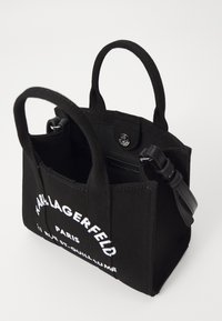 Mustakankainen käsilaukku, jossa on kaksi lyhyttä kahvaa, vetoketjulla suljettava ja nahkakaulus. Edessä on valkoista tulostettua tekstiä ja napin napsautus.