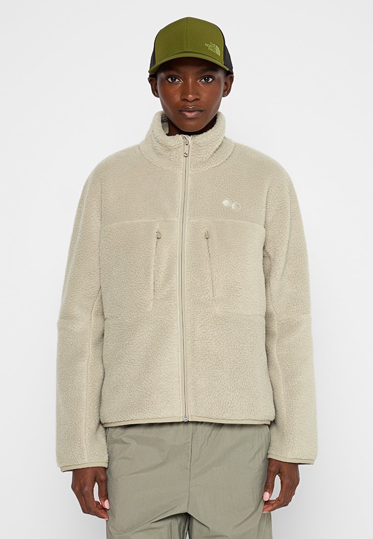 Person in einer hellbeigen Fleecejacke mit durchgehendem Reißverschluss und Brusttaschen, olivgrünen Hosen und einer olivgrünen Kappe von The North Face vor einem schlichten weißen Hintergrund.
