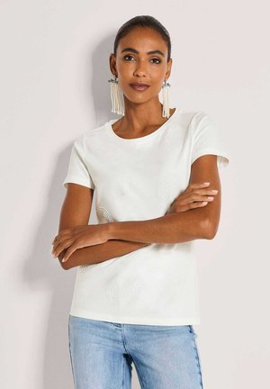 Vrouw met donker haar in een wit topje met korte mouwen en patroon en een lichtblauwe spijkerbroek, draagt lange kralenoorbellen, staat met gekruiste armen.