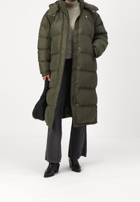 Olivgrön pufferjacka med huva, quiltad design, dragkedja fram och tryckknappar, bärs över en grå tröja och svarta vida byxor.