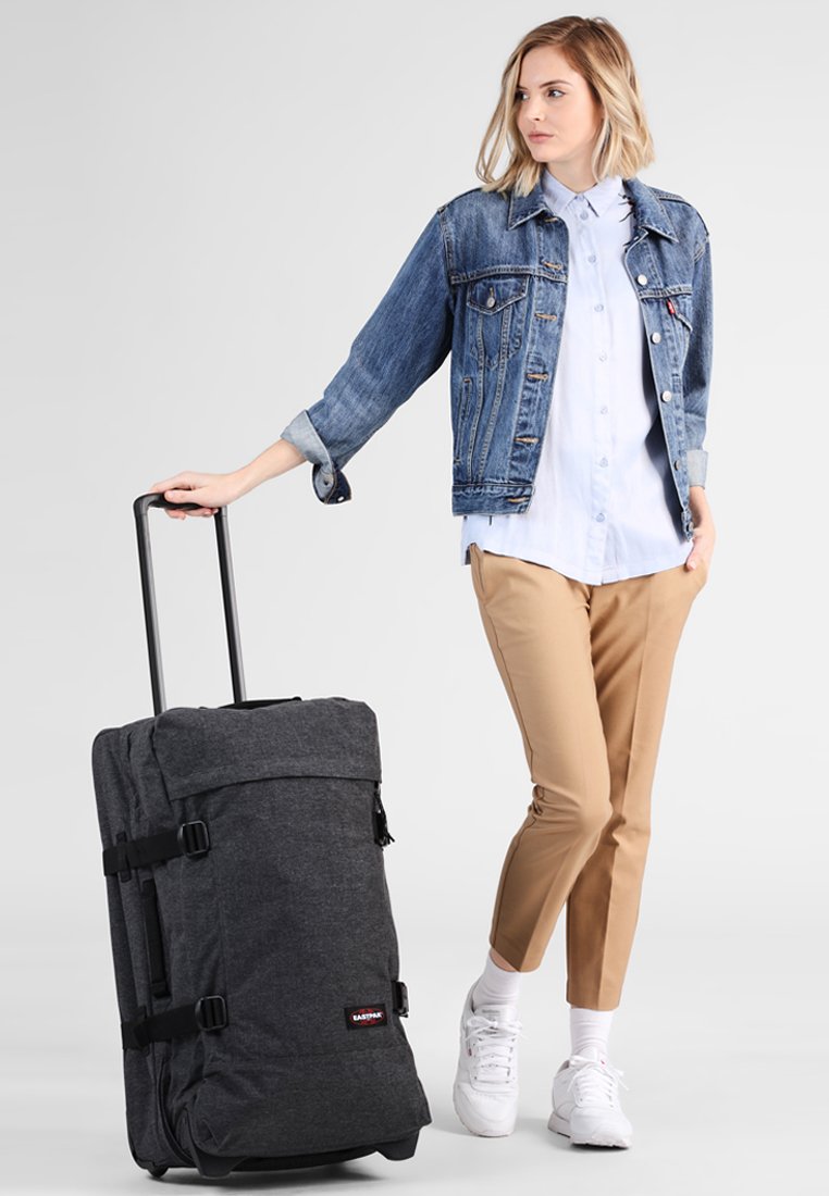 Borsa da viaggio nera con ruote, texture in tessuto, spalline regolabili e maniglia laterale. Modello in giacca di jeans e pantaloni beige si trova accanto ad essa.