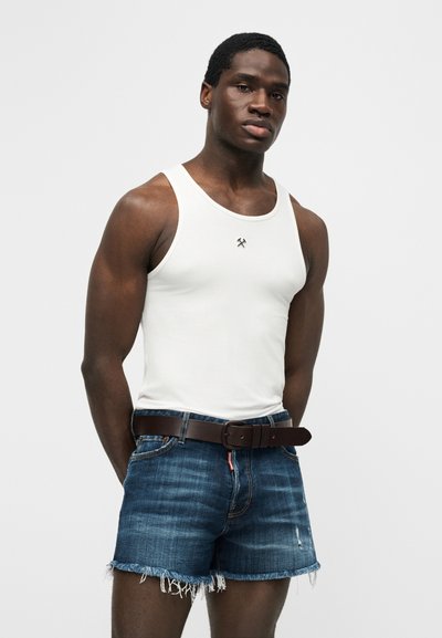 Άνδρας που φοράει λευκό αμάνικο μπλουζάκι με μικρό ανάποδα σταυρωμένο λογότυπο με σφυριά, σκούρο ζωνάρι και σκισμένο μπλε τζιν σορτς, μπροστά σε απλό φόντο.