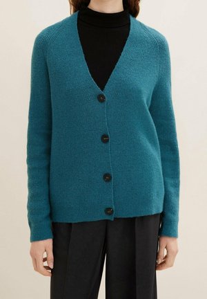 Femme portant un cardigan boutonné bleu sarcelle sur un col roulé noir et un pantalon noir, debout devant un fond beige uni.