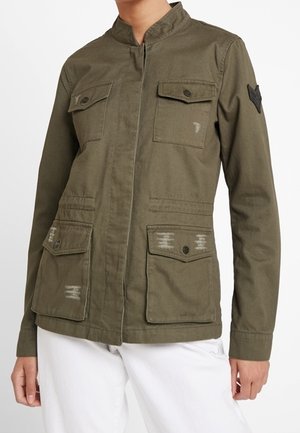 Oliwkowo-zielona kurtka w stylu militarnym z wysokim kołnierzem, klapowanymi kieszeniami na piersi i w biodrach, noszona z białymi spodniami, stojąca na białym tle.