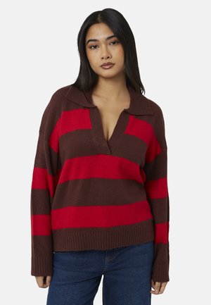 Junge Frau mit langen dunklen Haaren, die einen braun und rot gestreiften Pullover und blaue Jeans trägt, steht vor einem schlichten weißen Hintergrund.