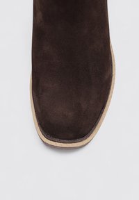 Botín de ante marrón hasta el tobillo con punta redonda, suela de goma beige y costuras visibles a lo largo del borde. Textura suave y diseño minimalista.