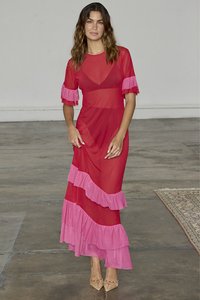 Transparent röd maxiklänning med lager av rosa volanger vid ärmar och nederkant, med en flytande silhuett och V-ringad design. Neutrala skor.