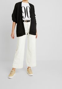 Schwarze Strickjacke mit offenem Vorderteil, kombiniert mit weißen, weit geschnittenen Hosen und beigen Sneakers. Das Outfit enthält ein grafisches T-Shirt mit großem "90"-Druck.