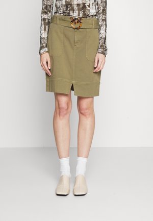 Femme portant une jupe verte olive longueur genou avec fente frontale, boucle de ceinture en écaille de tortue, haut à manches longues à motifs, chaussettes blanches et chaussures beiges.