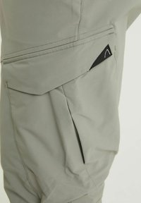 Poche de pantalon gris clair avec un rabat et une petite étiquette logo noire triangulaire sur le côté.