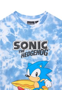 T-shirt in cotone blu con motivo tie-dye, caratterizzato da una grafica di "Sonic il porcospino", un personaggio dei cartoni animati in un vivace blu, una tavola da surf gialla e un testo audace.