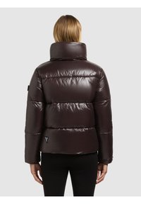 Kvinna som bär en glänsande mörkbrun vadderad jacka med hög krage, stående med ryggen mot en enfärgad grå bakgrund.