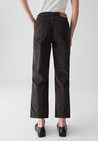 Persona che indossa pantaloni dritti marrone scuro e mocassini neri, in piedi contro uno sfondo chiaro uniforme, vista da dietro.