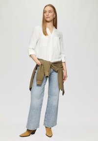 Chemise blanche à boutons avec deux poches sur la poitrine, associée à un jean bleu clair à jambe large et un pull olive noué. Des bottines marron viennent parfaire le look.