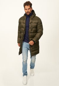 Olivgrüner Daunenmantel mit Kapuze, Reißverschluss und sichtbaren Nähten, getragen über einem marineblauen Pullover und hellblauen Jeans, kombiniert mit weißen Sneakern.