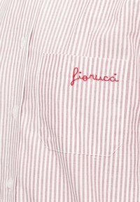 Bolso de camisa em tecido branco com finas riscas vermelhas verticais, bordado com texto em letras cursivas vermelhas "fiorucci," com a pala dos botões visível à esquerda.