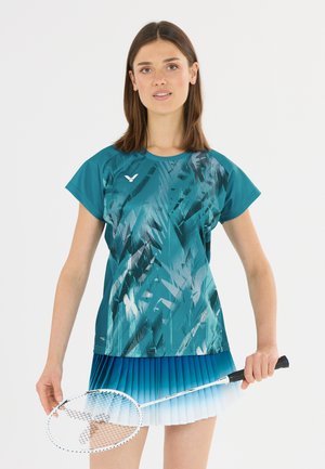 Ung kvinde i teal-mønstret sportstrøje og blå ombré plisseret nederdel, der holder en hvid badminton ketcher med sort håndtag.