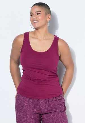 Smiling person with short hair wearing large hoop earrings, a sleeveless magenta top, and patterned loose magenta pants against a white background.