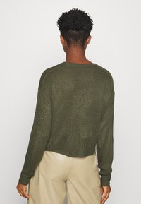 Olijfgroene gebreide trui met een relaxed fit, geribbelde manchetten en een cropped design, gecombineerd met beige high-waisted broeken.