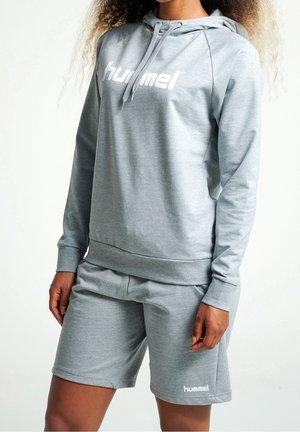 Frau trägt einen hellgrauen hummel Hoodie und passende Shorts, steht vor einem schlichten weißen Hintergrund.