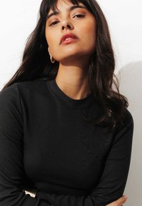 T-shirt noir à rayures côtelées à manches longues avec un col rond, présentant une texture lisse et une coupe ajustée. Le modèle affiche un maquillage discret et des accessoires minimalistes.