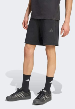 Osoba ubrana w czarne spodenki Adidas, czarne skarpetki Adidas oraz czarne buty sportowe Adidas, stojąca na jasnej powierzchni.