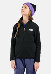 Pullover in pile nero con zip a metà, caratterizzato da un piccolo patch con logo. Abbinato a pantaloni blu, il modello indossa un berretto viola e nero.