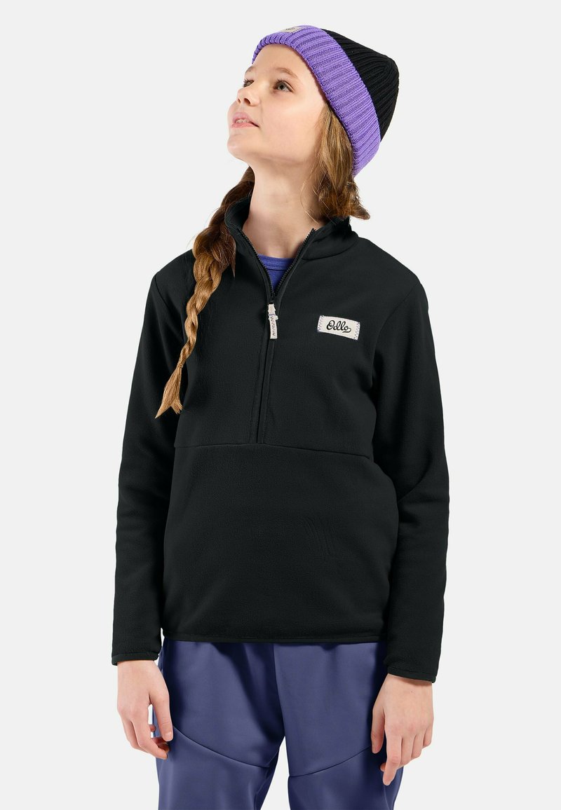 Pullover in pile nero con zip a metà, caratterizzato da un piccolo patch con logo. Abbinato a pantaloni blu, il modello indossa un berretto viola e nero.