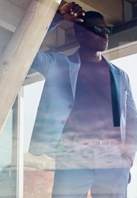 Lichtblauwe blazer met een slim fit, gedragen over een donker shirt. Model met donkere zonnebril, leunend tegen een houten paal. Textured riem zichtbaar.