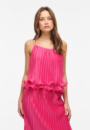 Femeie purtând o rochie fucsia, fără mâneci, plisată, cu straturi volanate în talie, stând pe un fundal alb simplu.